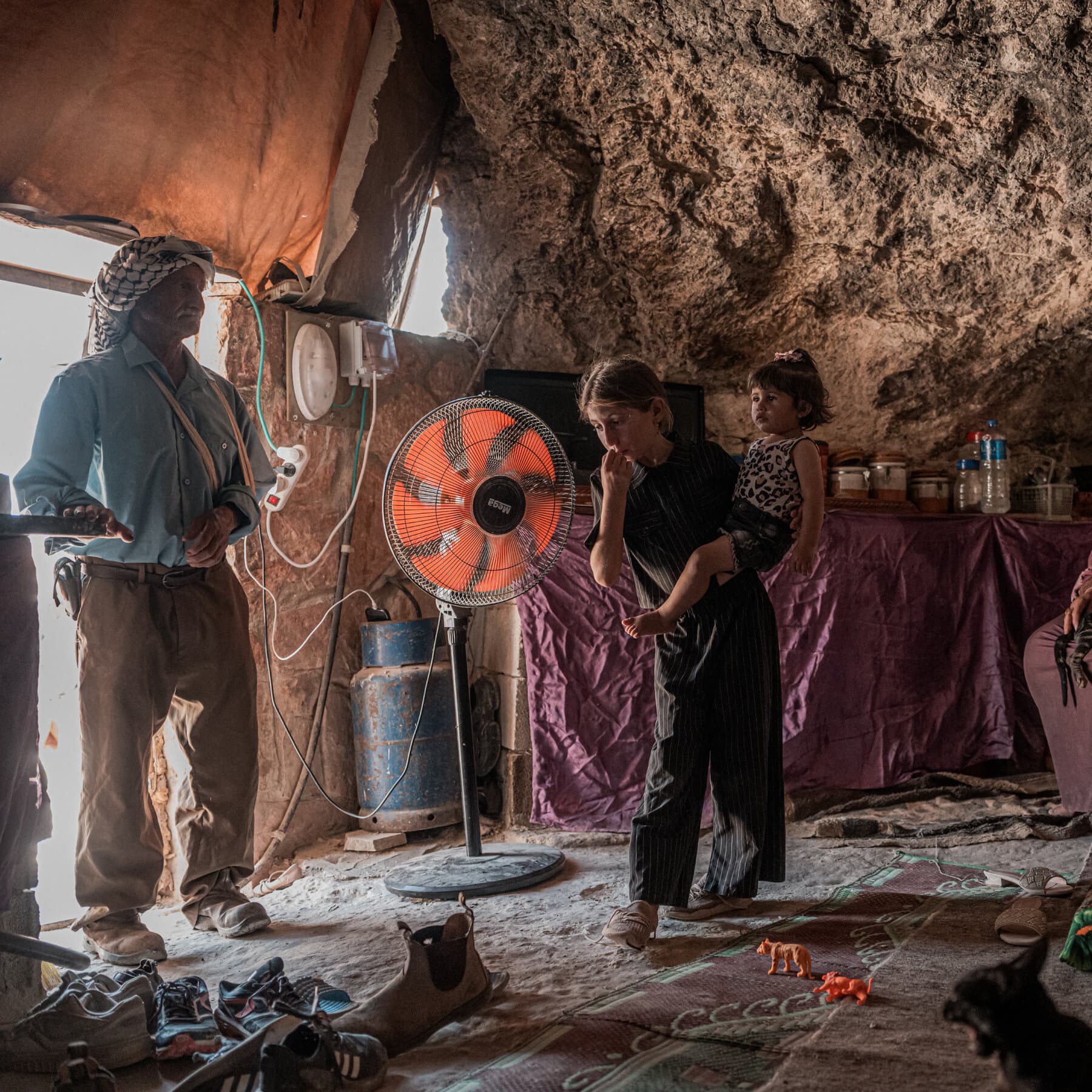 A desolate and dusty landscape in the West Bank featuring an abandoned stone house with a simple, worn-out appearance. In the foreground, a few left-behind personal items like a broken chair or a child's toy lie in the dirt. In the background, on a hill, shadowy figures of settlers are watching, silhouetted against a harsh, bright sky. The style should be gritty and realistic, capturing a sense of loneliness and defeat, similar to a high-contrast photojournalism piece.