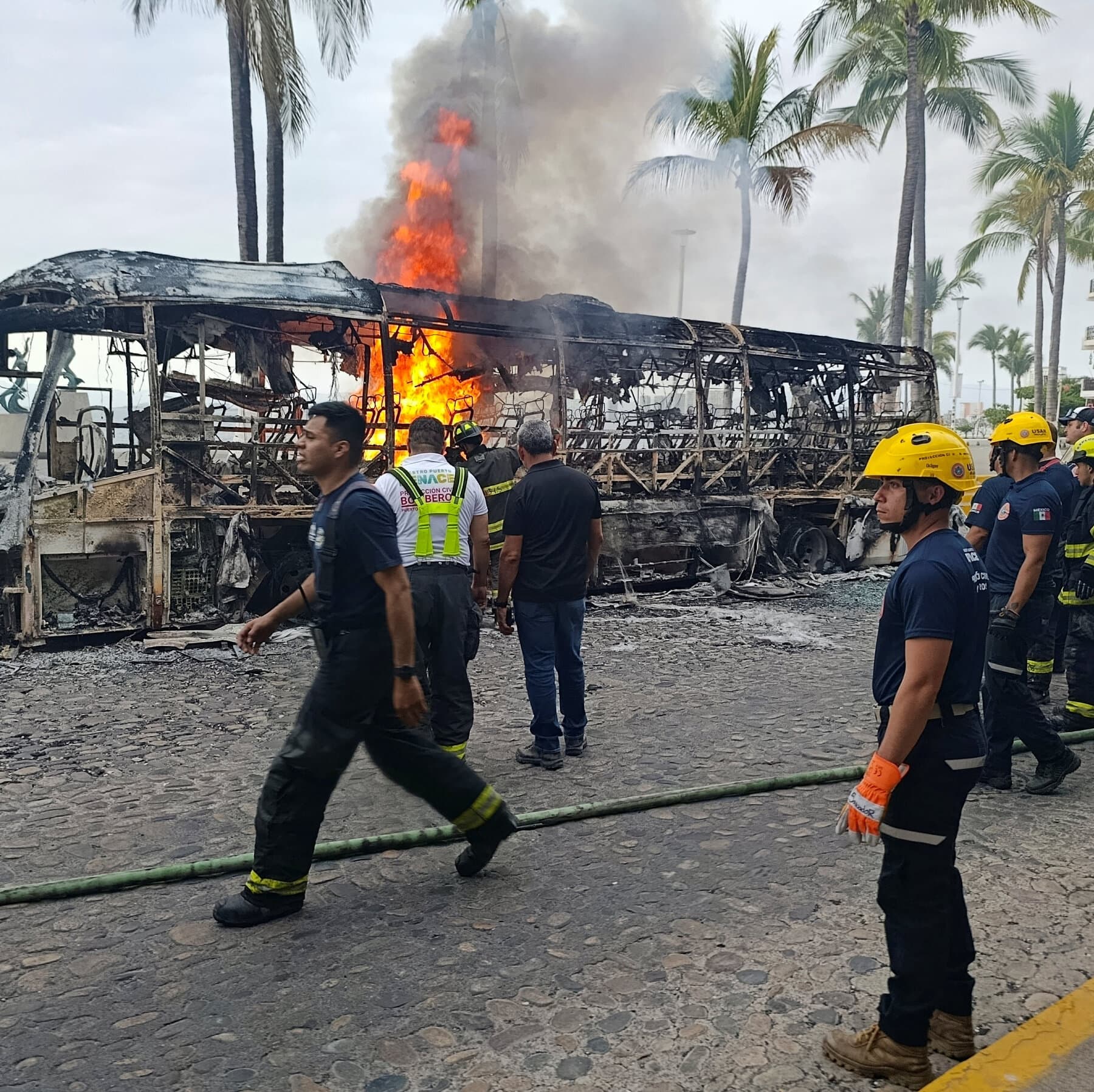 A split composition: on the left, a bright, sunny view of a beach resort with a colorful cocktail in the foreground; on the right, a dark, gritty street scene with a burning bus blocking the road and smoke rising into the sky. High contrast between the two worlds.