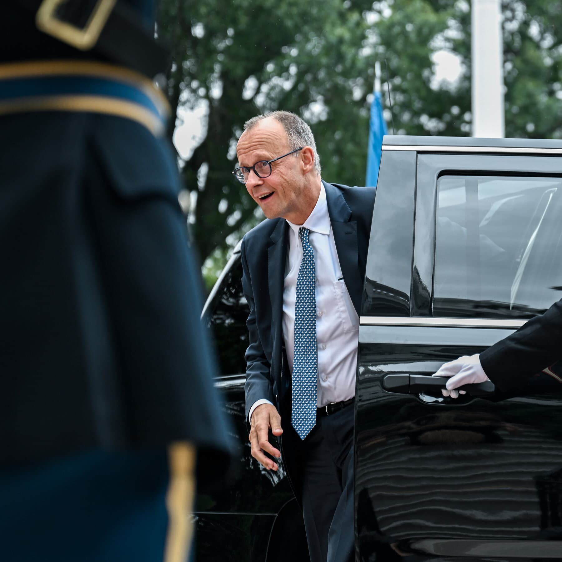 A gritty, cynical political cartoon style illustration. A small, nervous man in a German suit sweating profusely while balancing on a thin, fraying rope over a dark abyss. On one side of the cliff, a large, shadowy dragon waits. On the other side, a loud, orange-tinted storm cloud with a tie screams. The atmosphere is gloomy and tense. No text.