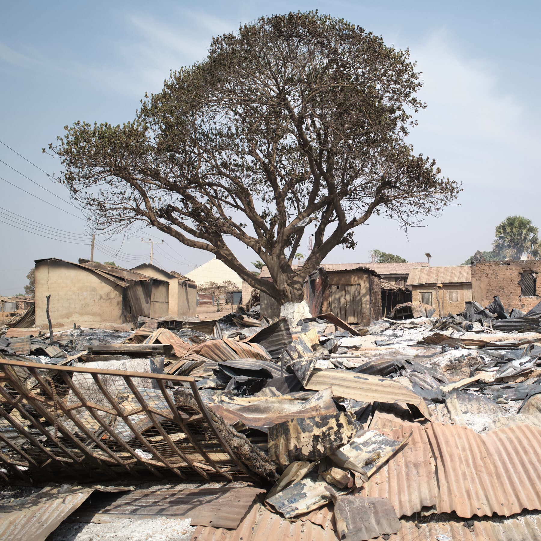 A highly satirical, gritty illustration in a political cartoon style. In the foreground, a giant, sleeping soldier in a pristine Nigerian military uniform is sitting in a comfortable armchair, cobwebs growing on his rifle. In the background, a small, weary-looking American cowboy figure is searching through tall grass with a magnifying glass. The setting is a chaotic, dusty landscape. The mood is cynical and dark.