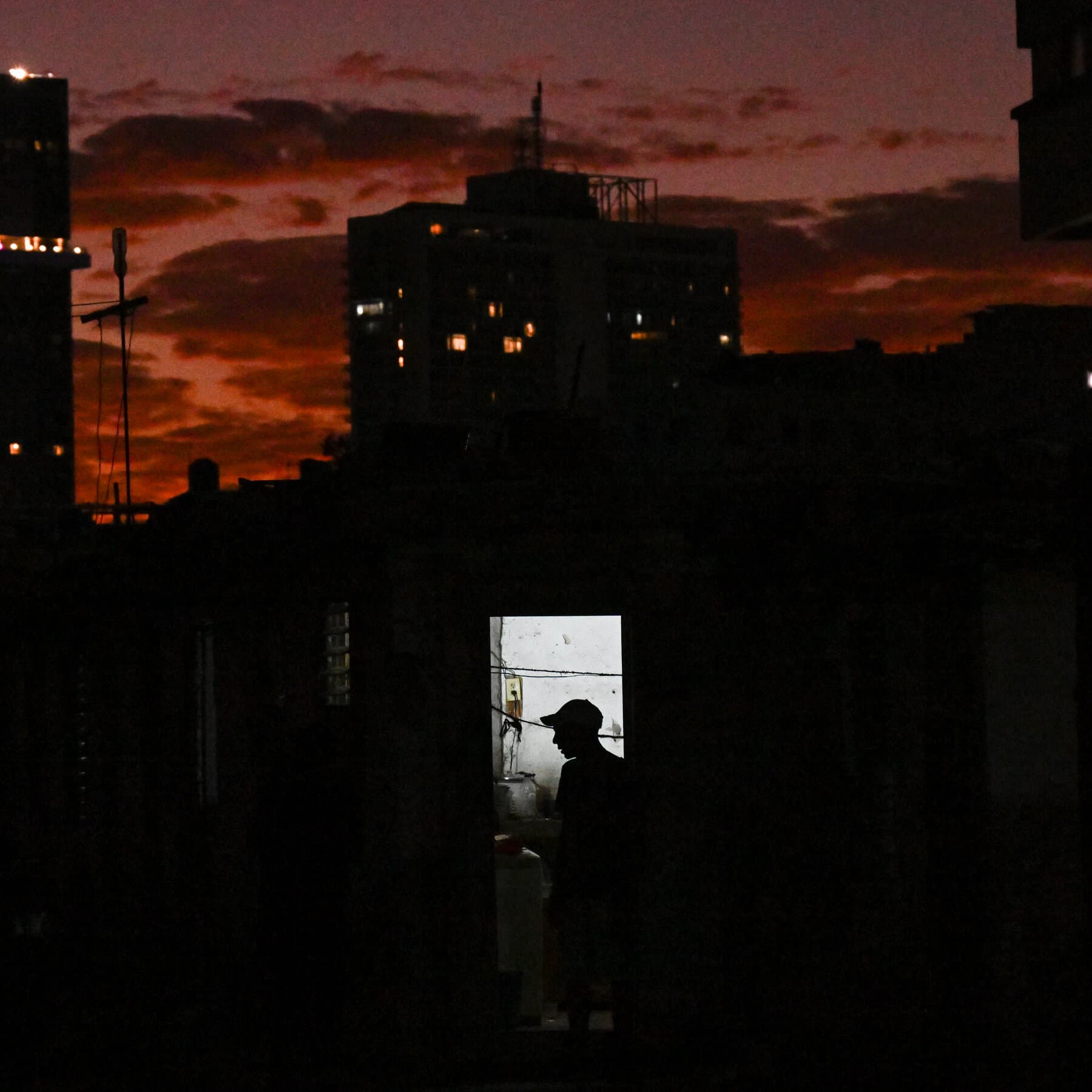 A gritty, cynical illustration in a political cartoon style. On one side, a large, orange-tinted hand in a suit sleeve is crushing a small, crumbling, colorful colonial building. On the other side, old men in green military fatigues are laughing while sitting on top of the rubble. In the middle, tiny, nondescript people are looking tired and empty-handed. The background is a stormy, grey sky over a tropical ocean.