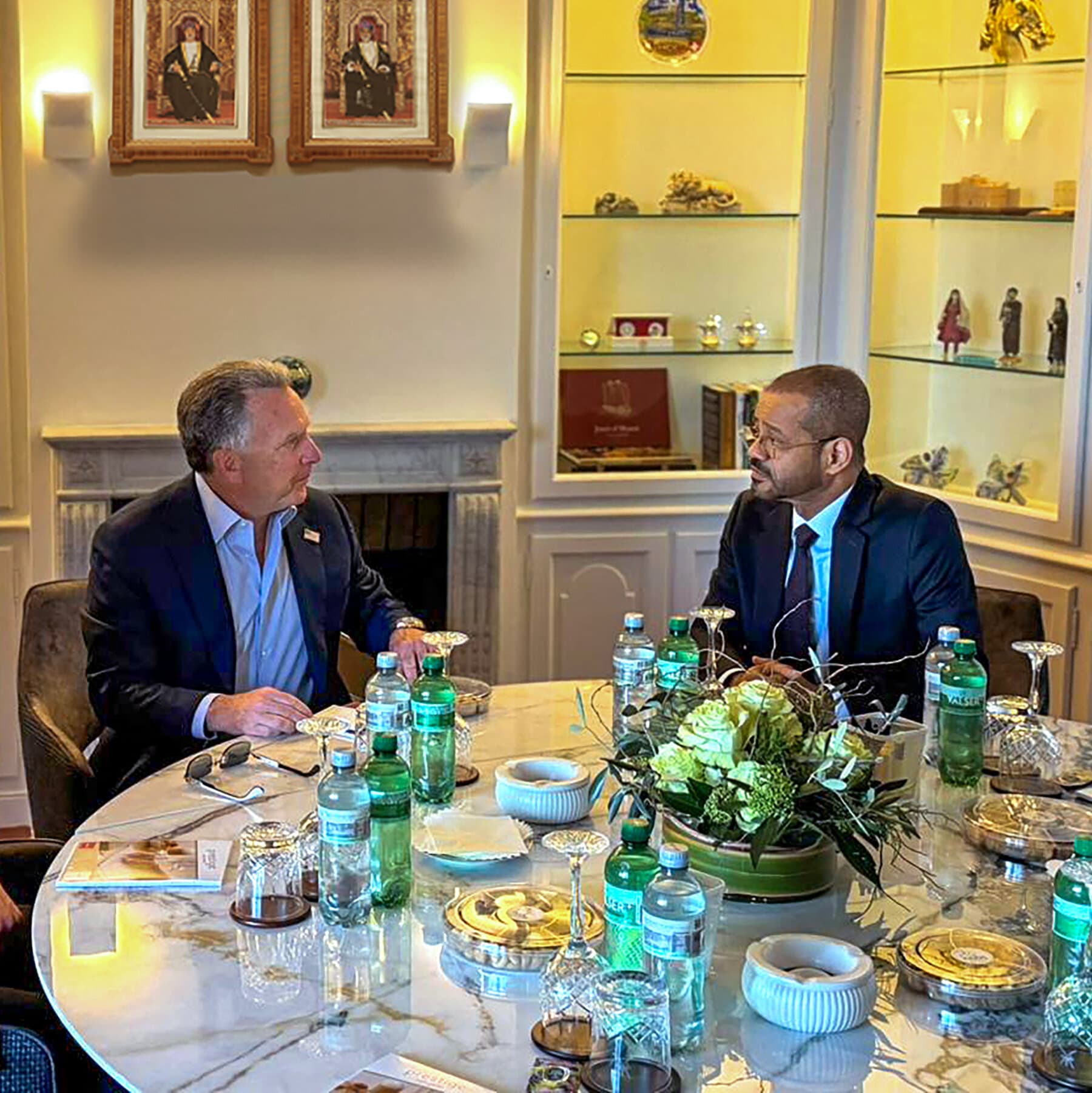 A surreal, dark political cartoon style image. A poker table sitting on top of a ticking bomb. On one side, a frantic, messy orange wig representing the US. On the other, a stern, dark turban. They are playing cards, but the cards are blank white rectangles. Smoke fills the room, and in the background, toy soldiers are melting.