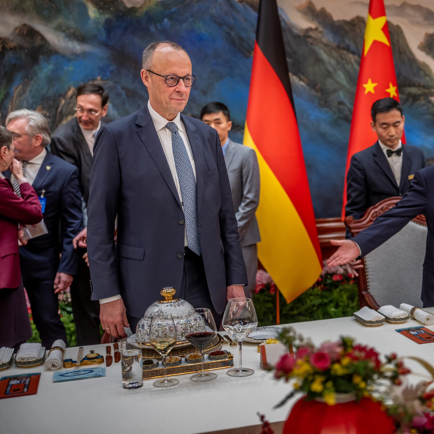 A gritty, high-contrast illustration of a small, tired-looking man in a suit trying to hold apart two massive, crushing walls. One wall is painted with an American flag pattern, the other with a Chinese flag pattern. The man looks exhausted. In the foreground, blurry silhouettes of fat cats in tuxedos are laughing and counting money, ignoring the man being crushed. The style should be dark, cynical political cartoon art.