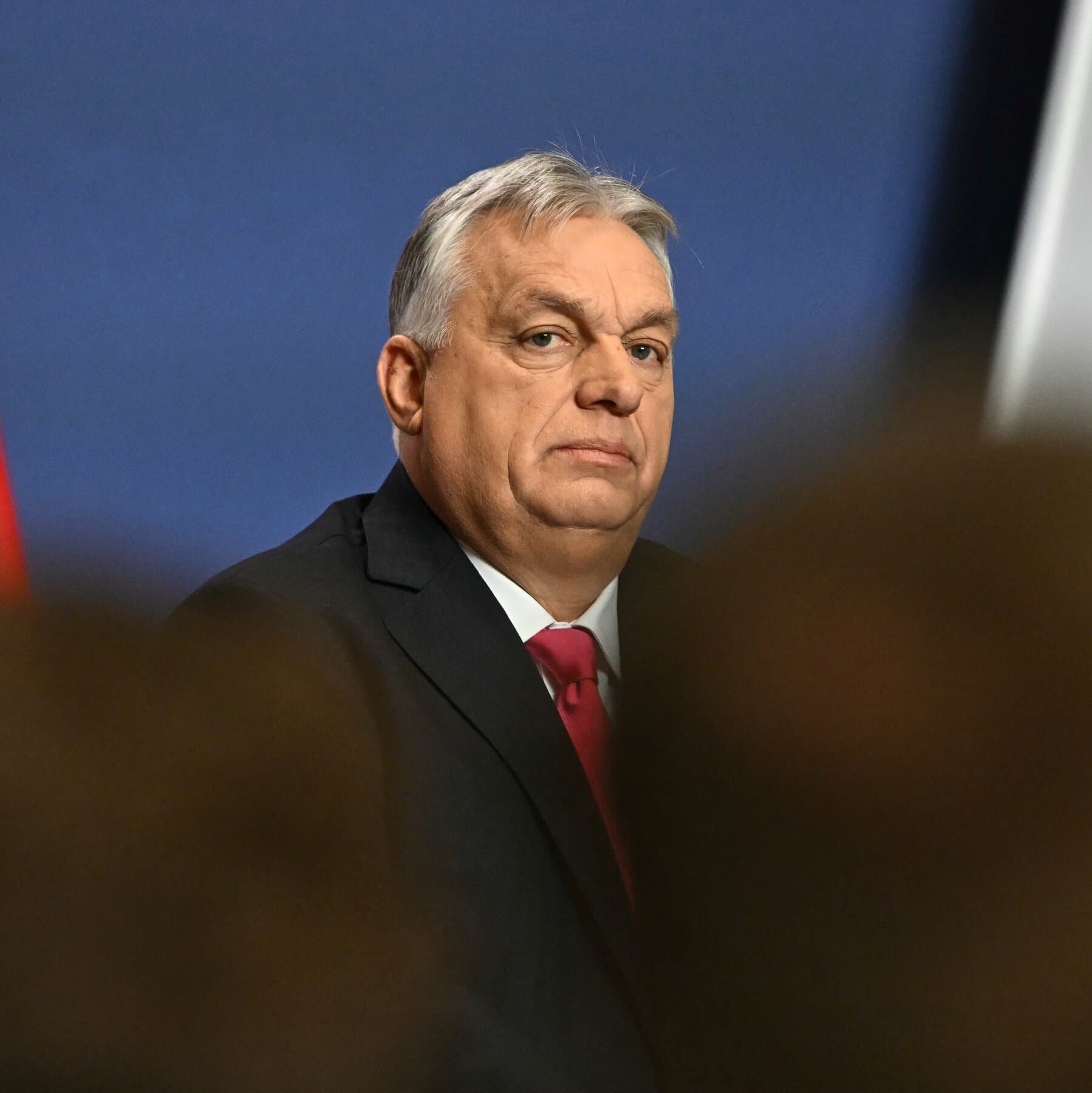 A satirical political cartoon style illustration showing a small, grumpy man in a suit standing alone in a large, blue European parliament chamber. He is holding a giant red stop sign. Surrounding him are other politicians in suits who look tired, annoyed, and exasperated. In the background, stacks of money are locked inside a cage. The lighting is dramatic and moody, emphasizing the isolation of the man with the stop sign. High contrast, editorial style.