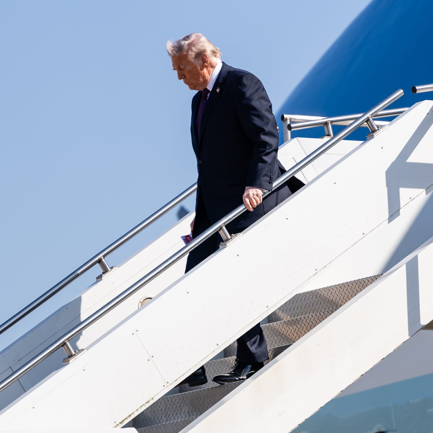 A gritty, desaturated photo of three pairs of empty, dusty combat boots arranged in a row on cracked desert earth. In the background, out of focus, is a blurred image of a suit and tie on a podium. The lighting is harsh and unforgiving. No faces visible. Mood is bleak and cynical.