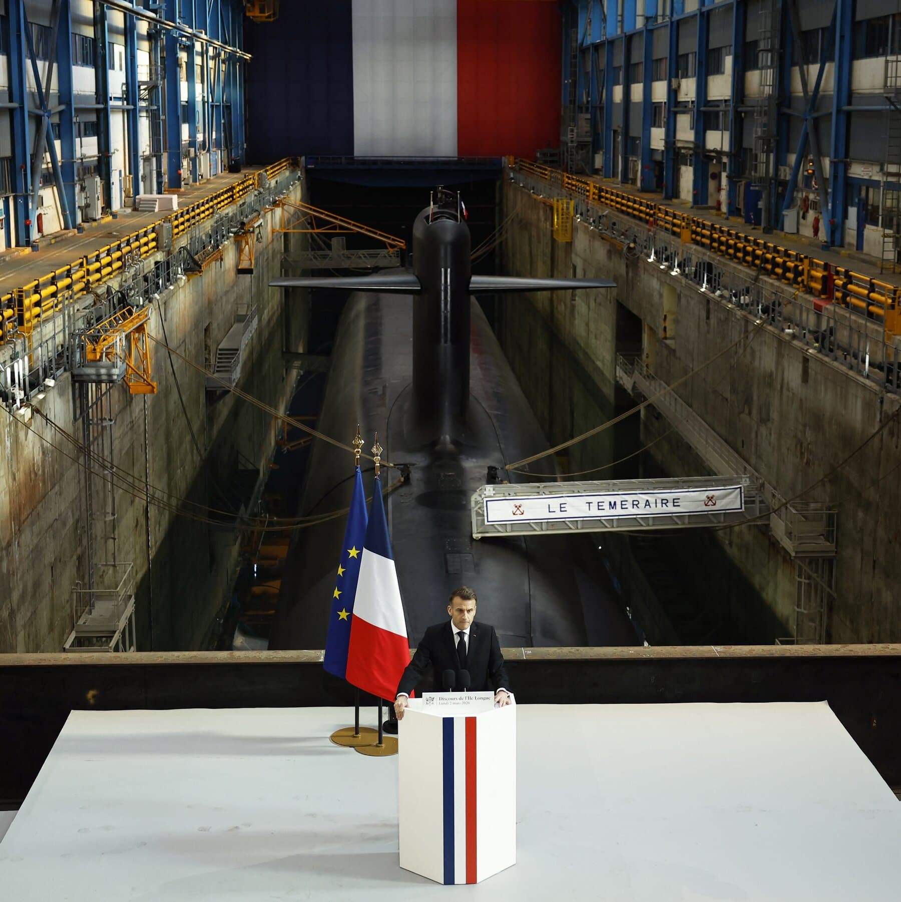 A cynical political cartoon of Emmanuel Macron in a sharp suit, standing on a podium shaped like a nuclear missile. He is looking down at a crowd of tiny, confused people while holding a small toy globe. The background is a dark, stormy sky over a map of Europe. The style is gritty and dark, with a sarcastic tone.