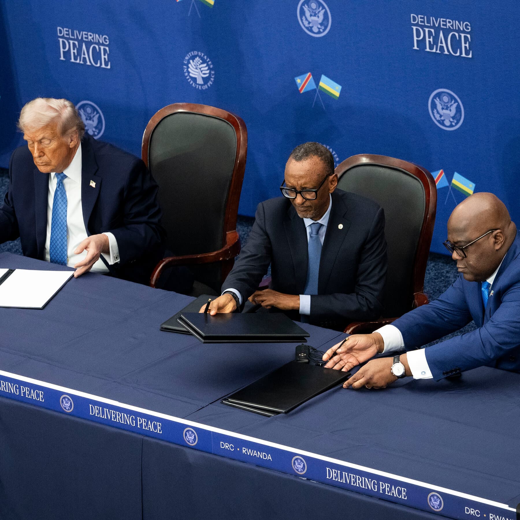 A conceptual illustration in a gritty, cynical style. On one side, a pristine, signed document labeled 'PEACE DEAL' resting on a mahogany desk. On the other side, a rugged, torn map of Rwanda being stamped with a heavy red ink stamp that reads 'SANCTIONED'. The lighting is dim and moody, emphasizing the disconnect between the office and the reality.