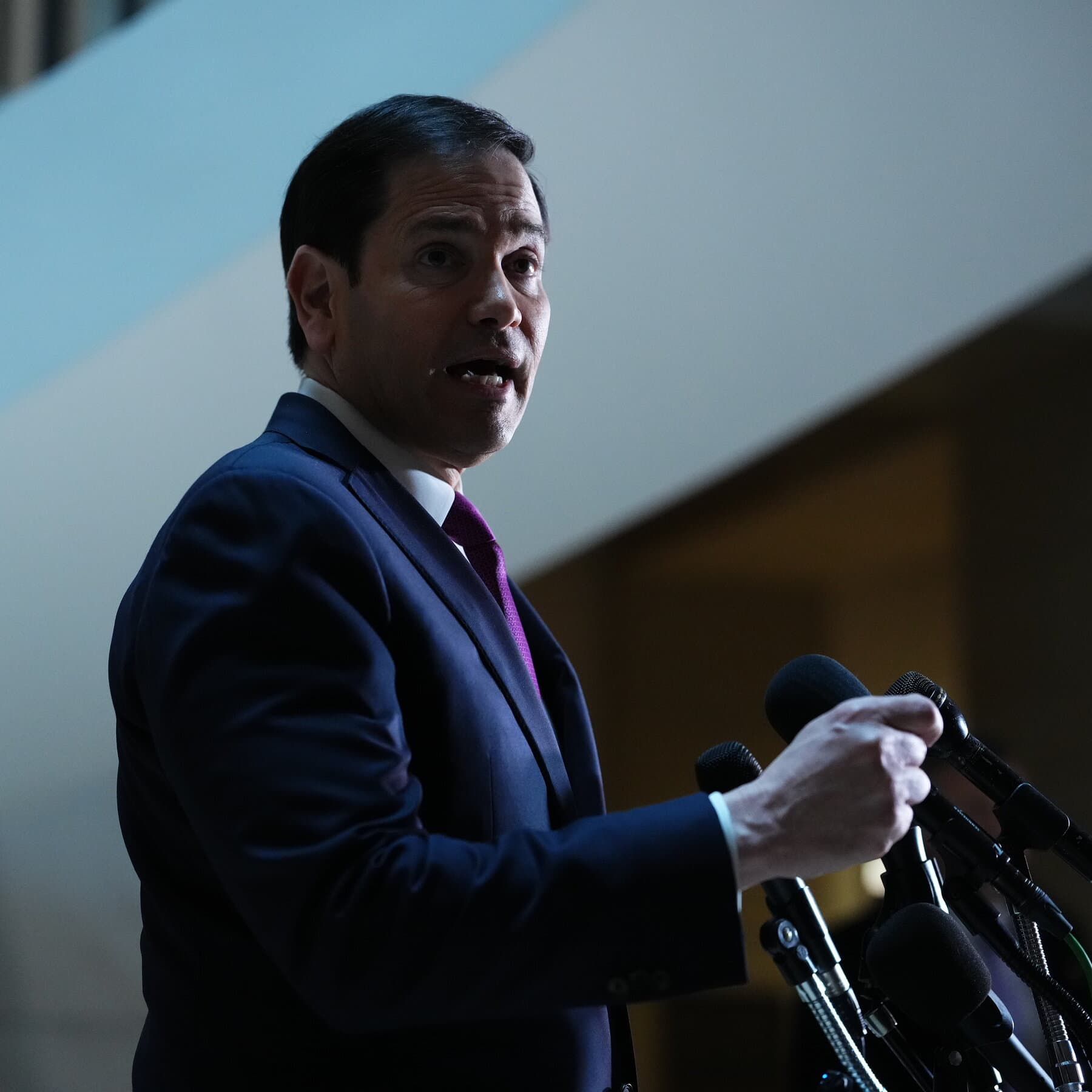 A political figure in a sharp suit standing behind a podium made of old, crumbling stone. The background is a theatrical stage with dark red curtains that are slightly on fire. On the floor are toy missiles and confusing maps. The lighting is dramatic and cynical, oil painting style.