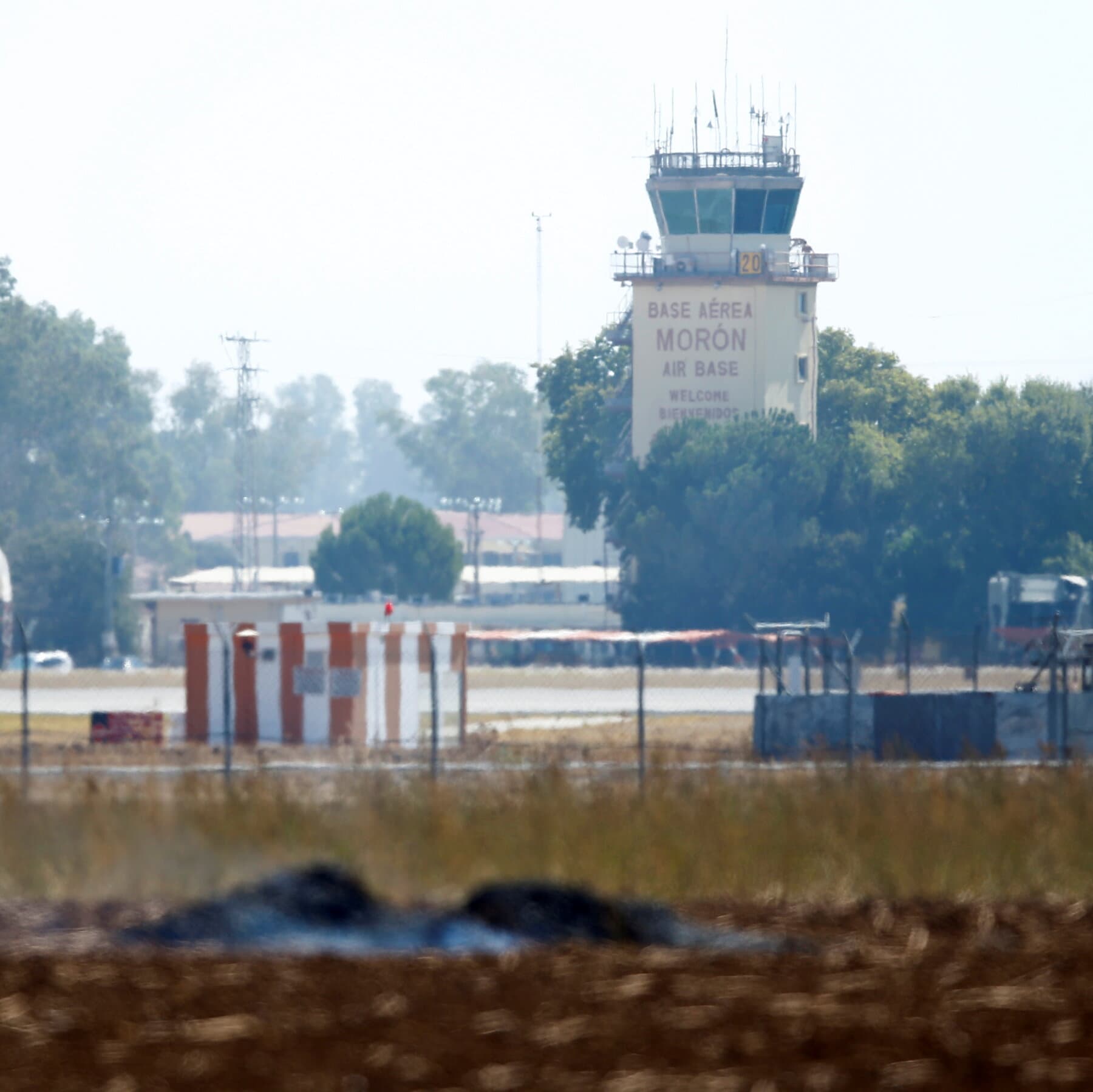 A gritty, cynical editorial cartoon style illustration showing a rusted metal gate with a 'Do Not Enter' sign written in Spanish. On one side of the gate, a fat, angry general with a cigar and a US flag patch is kicking the fence. On the other side, a smug Spanish politician in a suit is holding a rulebook and laughing while looking at a mirror. The sky is grey and gloomy. No text bubbles.