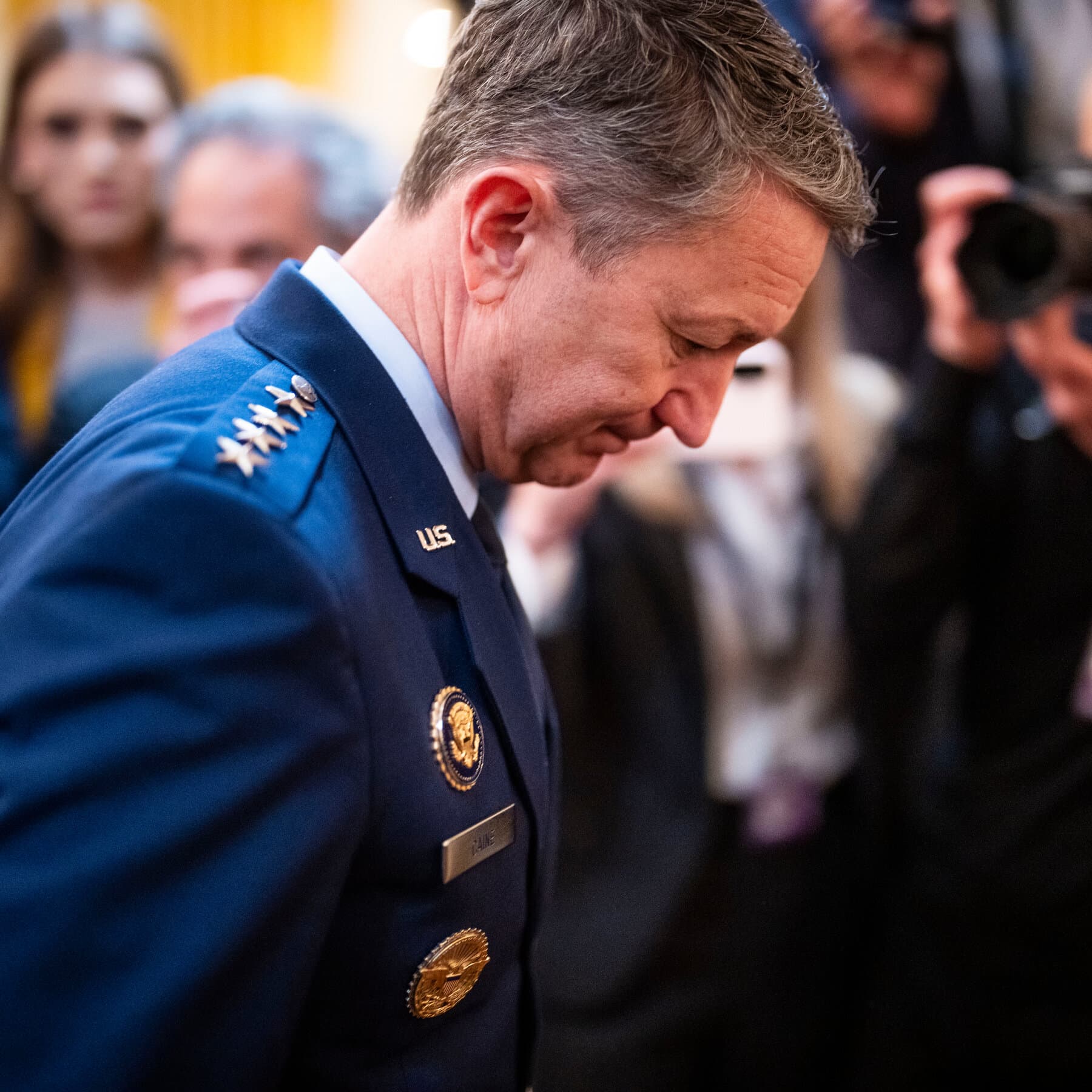 A gritty, high-contrast illustration in the style of a political cartoon or noir graphic novel. Focus on a vast, empty desert landscape under a harsh sun. In the foreground, six empty pairs of military boots lined up in the sand, half-buried. In the far distance, blurred silhouettes of men in business suits shaking hands. No text. Muted earth tones with a stark red sky.