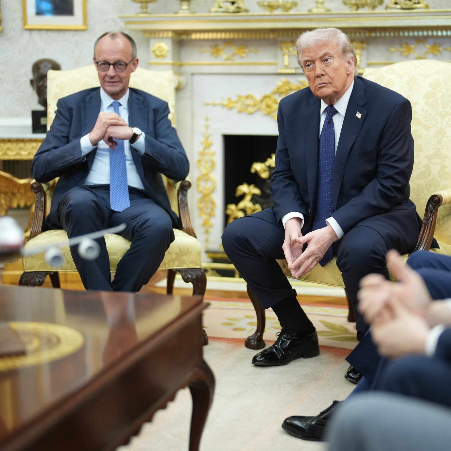 A gritty, stylized illustration of a chaotic Oval Office meeting. A large, loud figure in a suit gestures angrily at a map of Spain. A second figure, representing a German leader, sits stiffly in a chair, looking down at his hands, fading into the background wallpaper. The atmosphere is tense and cynical.