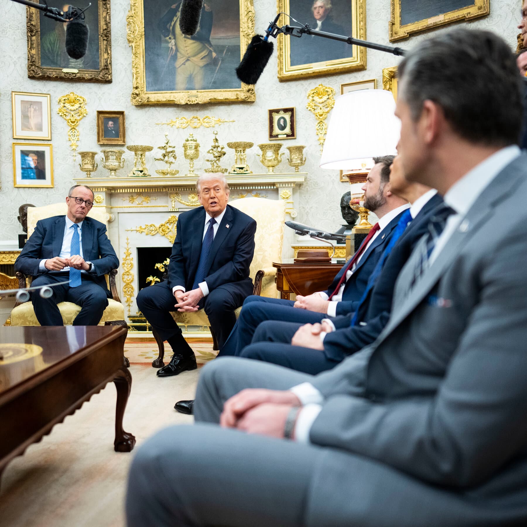 A conceptual illustration of Donald Trump standing on a tightrope, looking stressed and trying to balance. On one side of the rope below him is a crowd of angry supporters wearing red hats, shouting and pointing. On the other side is a chaotic scene of military tanks and desert conflict. The background is a dark, moody theater stage with a spotlight on the tightrope walker. The style should be high-contrast, satirical political art.