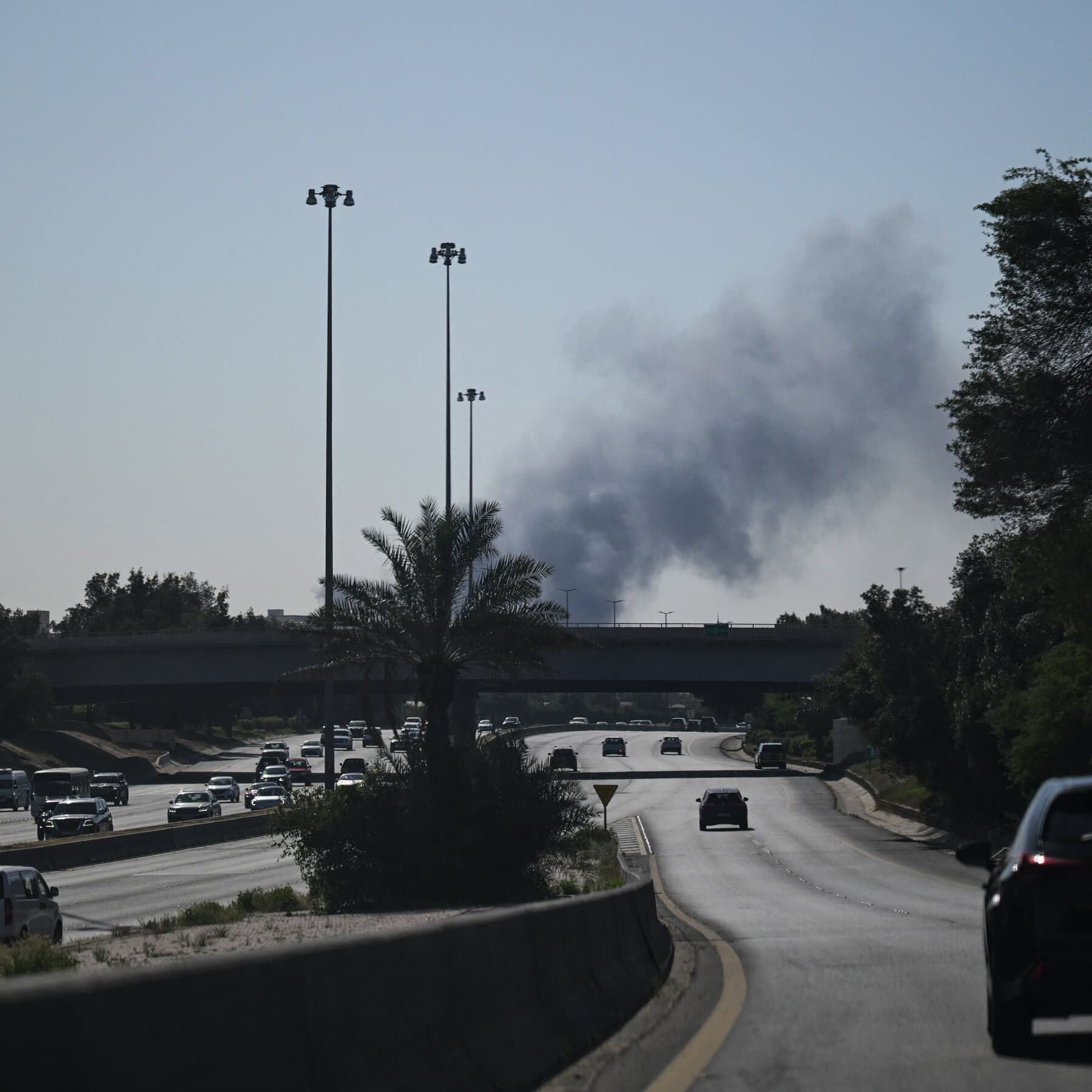 A moody, cynical illustration of a closed US embassy gate in a desert city at twilight. A heavy chain and padlock secures the gates. In the background, a modern skyline is silhouetted against a dusty, orange sky. A discarded newspaper blows across the empty street in the foreground. The atmosphere is abandoned and quiet.
