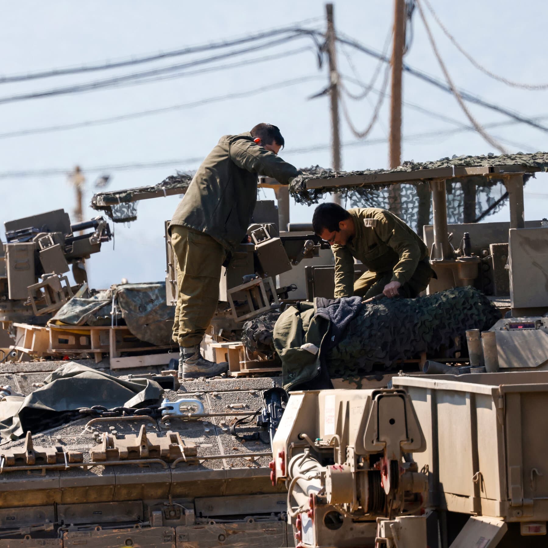 A gritty, high-contrast political cartoon style illustration. A giant, rusty meat grinder sits in the middle of a desert landscape. One side is painted with a blue flag, the other with a yellow and green flag. Both sides are feeding piles of money and small, faceless toy soldiers into the top. Out of the bottom comes red sludge that forms the shape of a map. In the background, fat men in suits are toasting with wine glasses filled with oil. The sky is dark and smoky.