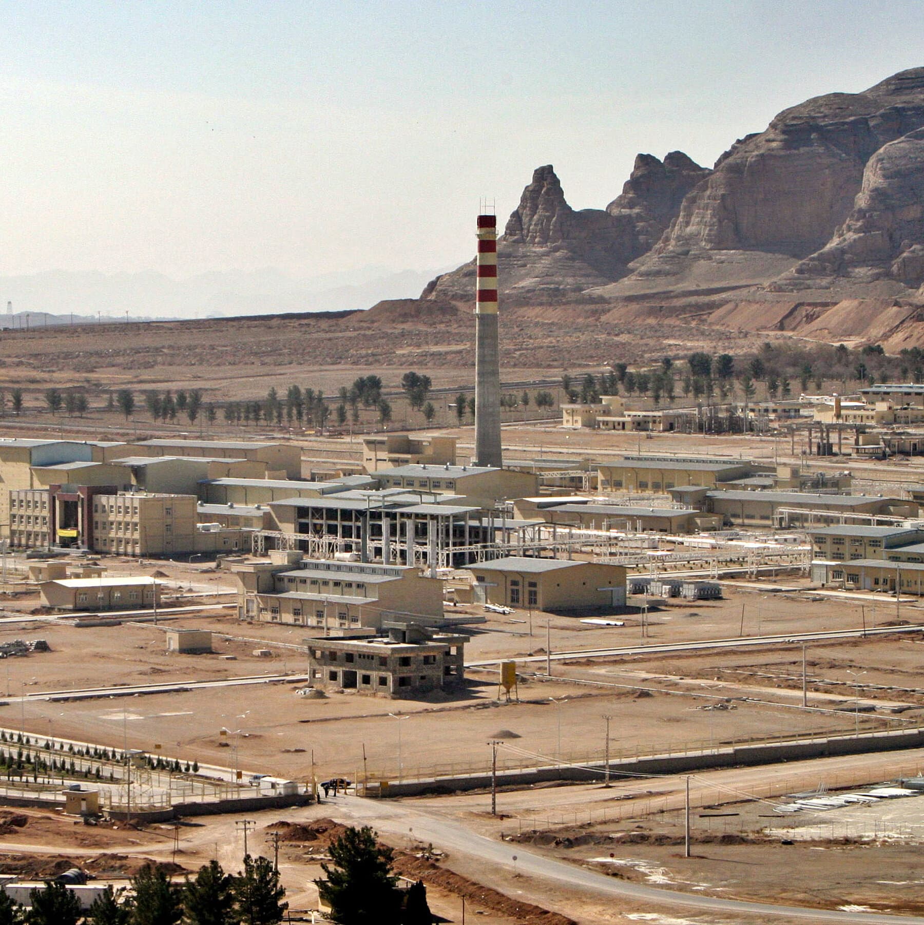 A gritty, high-contrast black and white image of a concrete industrial facility in the desert viewed through the circular lens of military binoculars. The image is grainy. A bright red digital overlay text in the center reads 'PAUSED'. The sky is bleak and overcast.