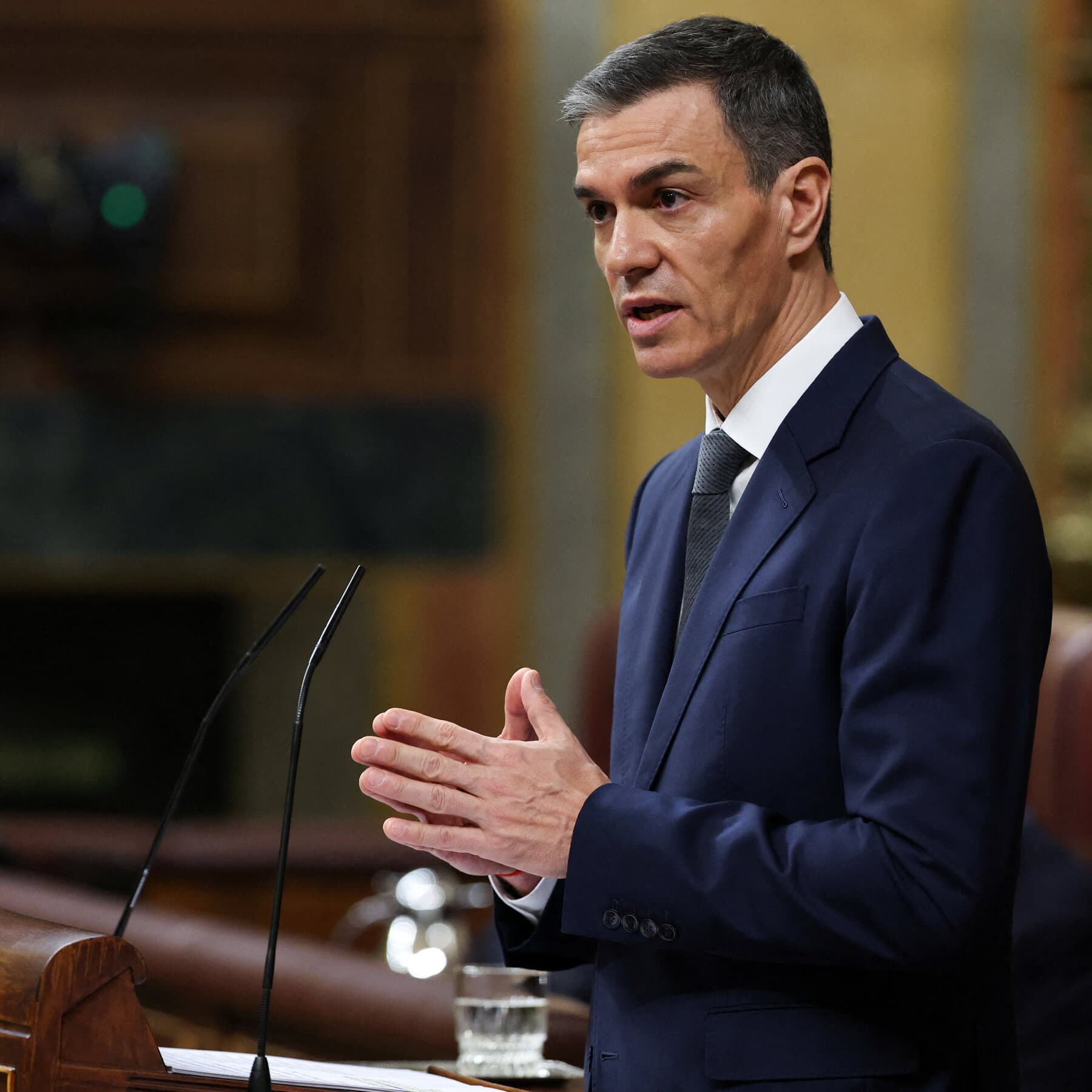 A satirical political cartoon style illustration. On the left, a figure resembling Pedro Sánchez stands on a small wooden crate, looking noble and defiant, holding a paper shield. On the right, a massive, looming shadow of a figure resembling Donald Trump casts a dark silhouette over him, holding a bag of money used as a weapon. The background is a chaotic, abstract map of the world in flames. High contrast, ink and watercolor style, moody and cynical atmosphere.