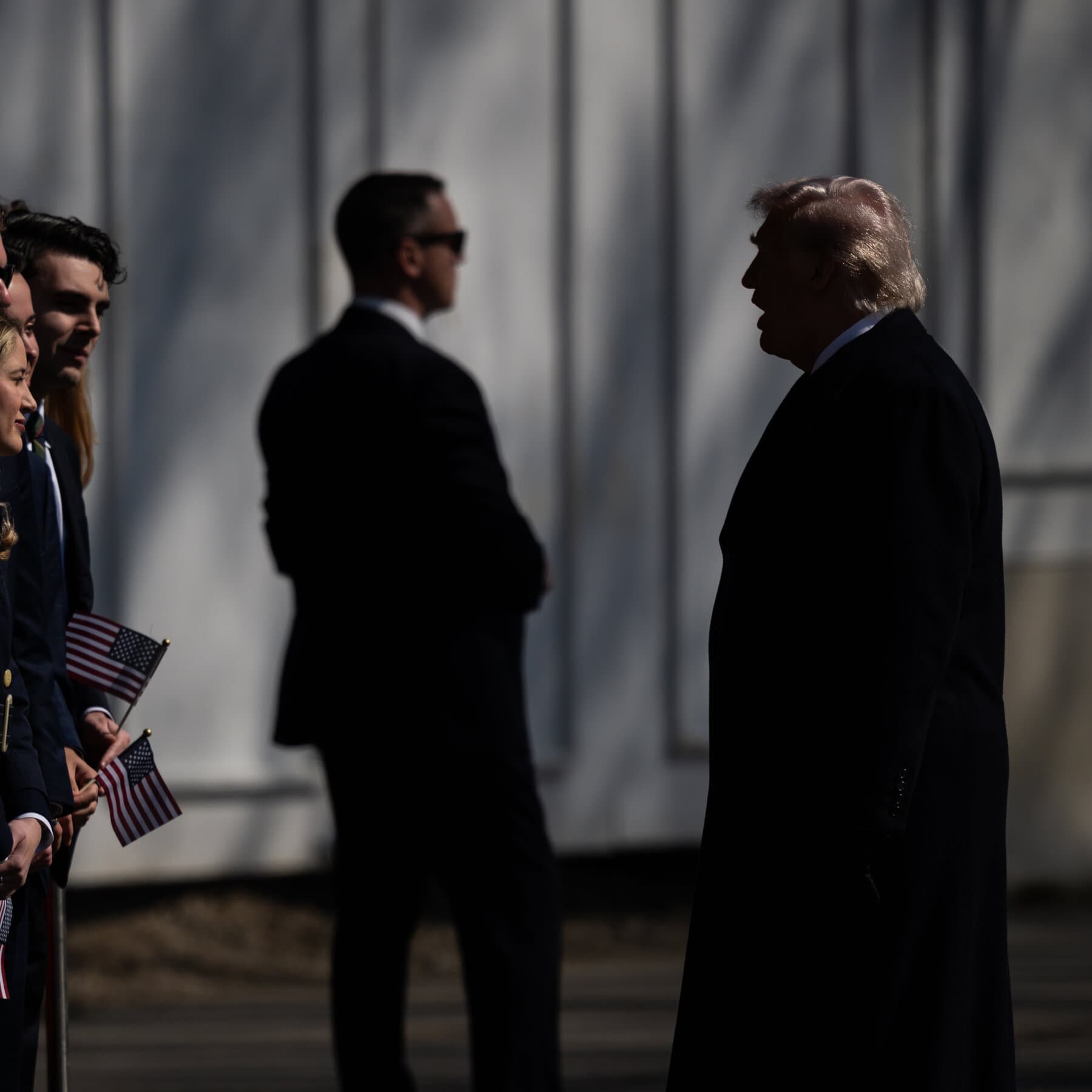 A gritty, noir-style courtroom sketch. A lone, shadowy figure sits in the witness box, gesturing with open hands as if pleading. In the background, looming large and abstract, is a dark map of Iran casting a shadow over the room. The atmosphere is cynical, dusty, and tired. The colors are muted grays and browns.