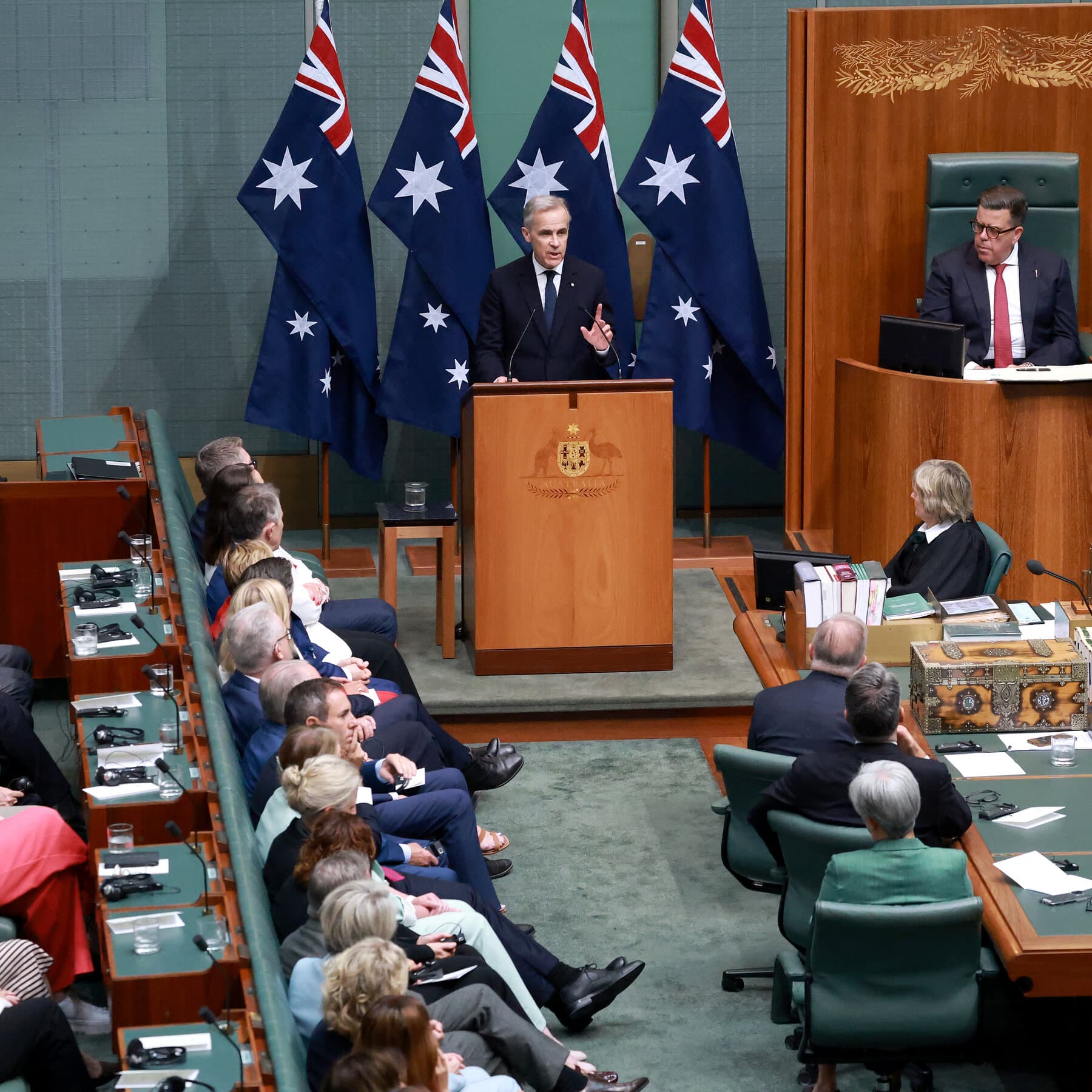 A conceptual editorial illustration showing two small, polite beavers and a kangaroo sitting at a tiny tea table in the middle of a wrestling ring, holding up a piece of paper that says 'Agenda', while giant shadowy wrestlers fight in the background.