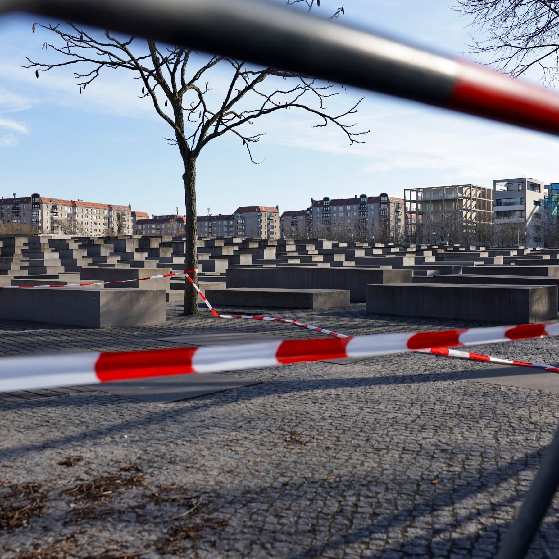 A moody, desaturated photograph of the Berlin Holocaust Memorial with its gray concrete steles rising like a maze. In the foreground, a single, sharp focus on an empty bench or step with a discarded tourist map, symbolizing the disruption of peace. The lighting should be overcast and gloomy, capturing a sense of cold, bureaucratic cynicism.