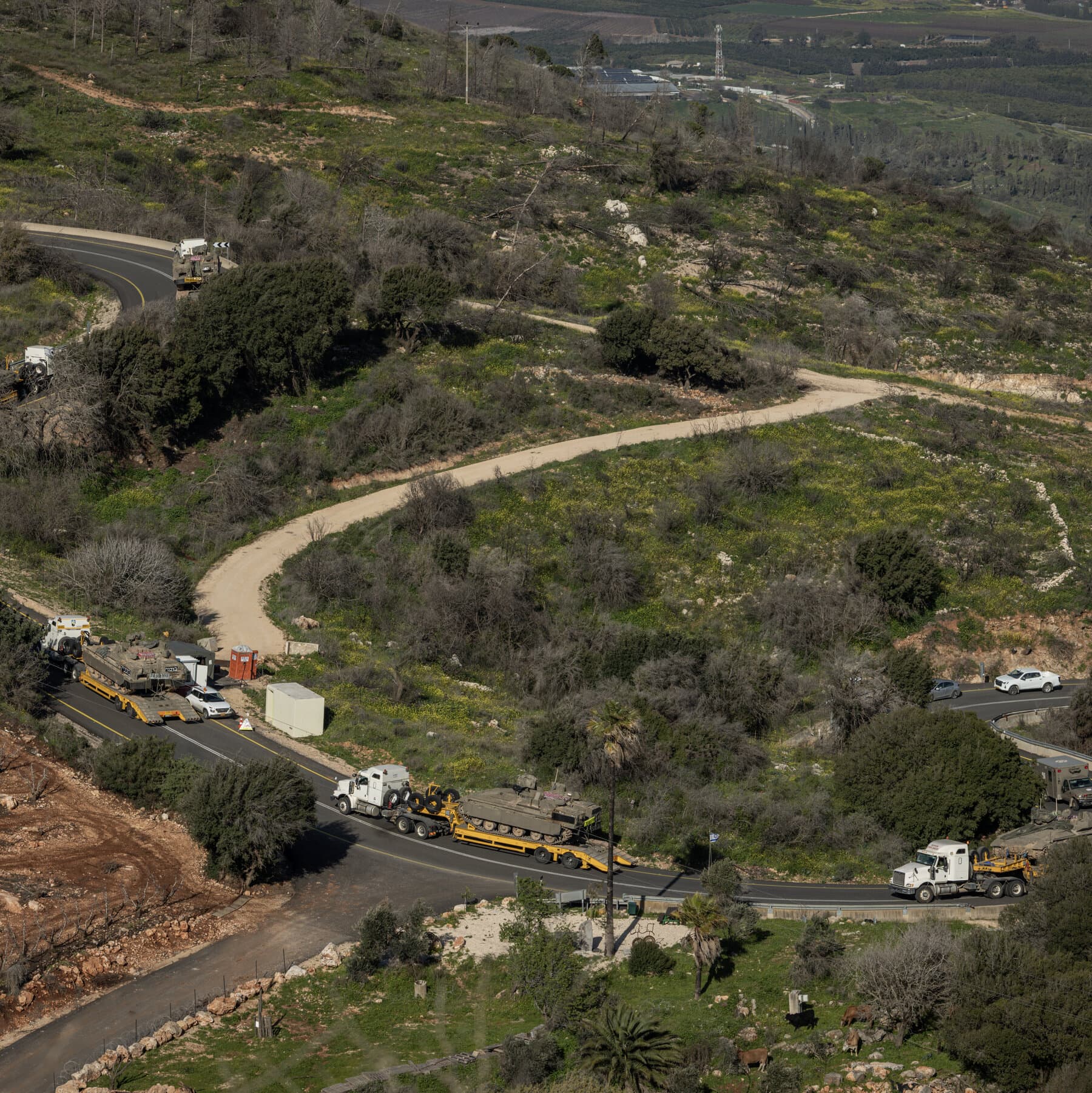 A gritty, black and white photojournalism style image. In the foreground, the muddy heavy treads of a tank are visible, focused and sharp. In the background, out of focus, a long line of armored vehicles stretches into a dusty, hazy horizon under a gloomy grey sky. No people visible, just the cold machinery of war massing in a desolate landscape. High contrast, cynical and somber mood.