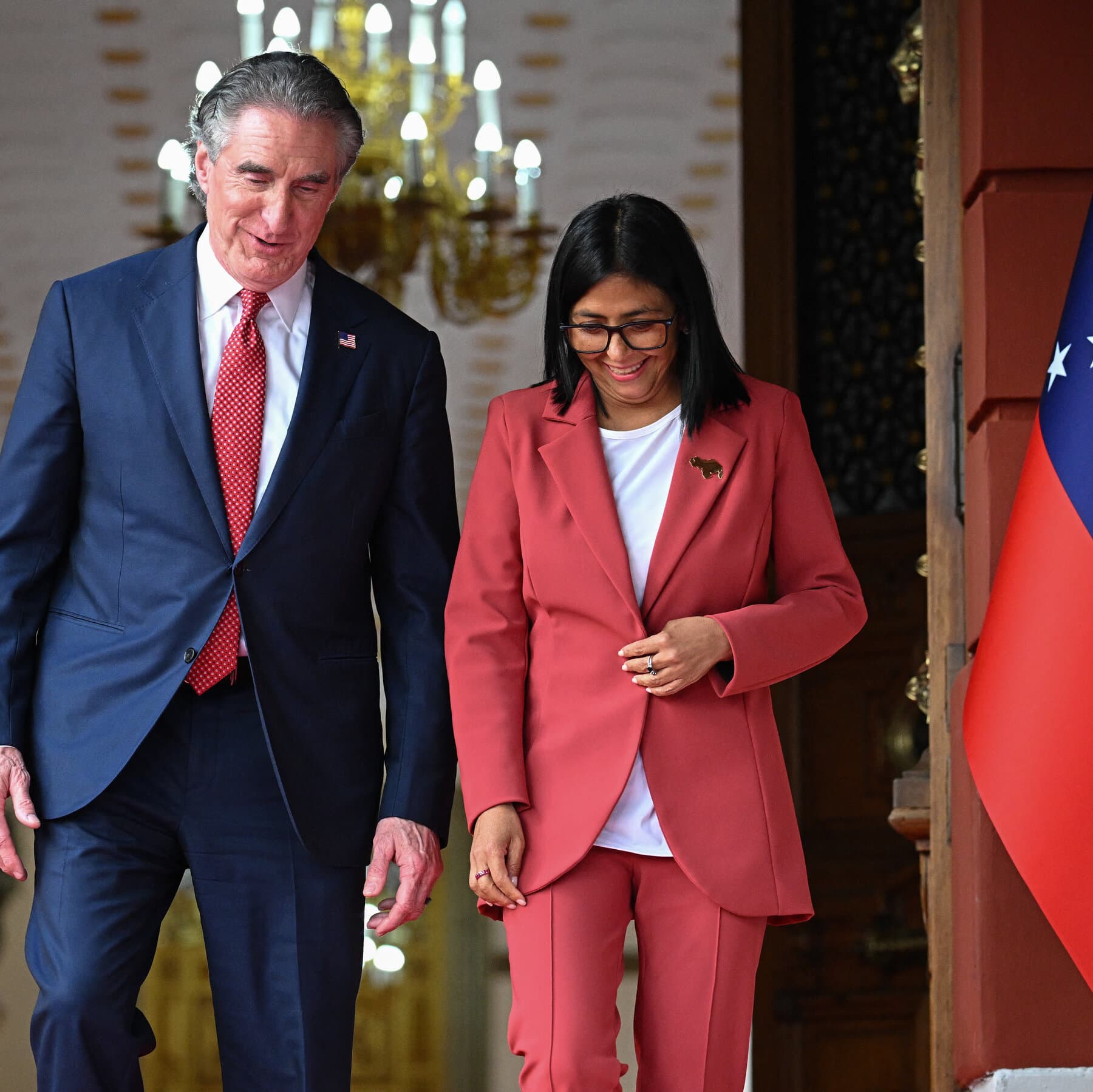 A satirical, high-contrast political illustration of a business suit sleeve with a hand reaching down from the top of the frame, squeezing a map of Venezuela. The map is cracking, leaking gold liquid and black oil. The background is a dark, moody grey. No text.