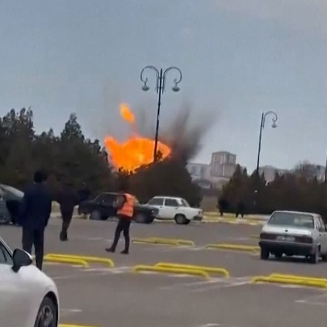 A desolate, muddy landscape with a smoking, crashed gray drone in the foreground. In the background, a blurred barbed wire fence. Overcast, gloomy sky. Realistic photojournalism style.
