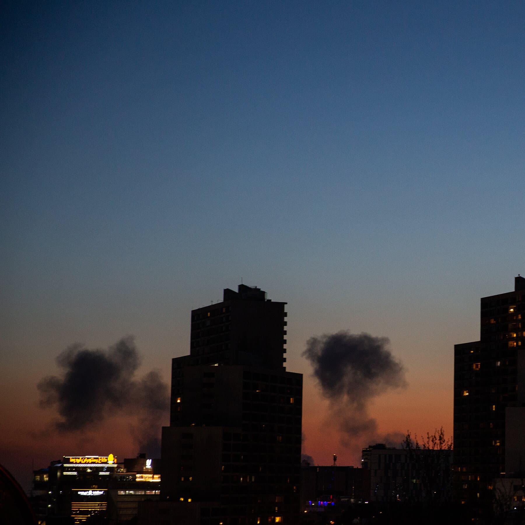 A satirical illustration in a gritty, high-contrast style. In the foreground, a large, exaggerated silhouette of a politician in a suit stands behind a podium, shouting into a microphone. The microphone cord transforms into a lit fuse. In the background, a split scene shows a map of the Middle East crumbling like a dry cookie, with stylized smoke plumes rising from Tehran and Beirut. The colors should be muted grays, dark reds, and sickly yellows to convey toxicity and cynicism.