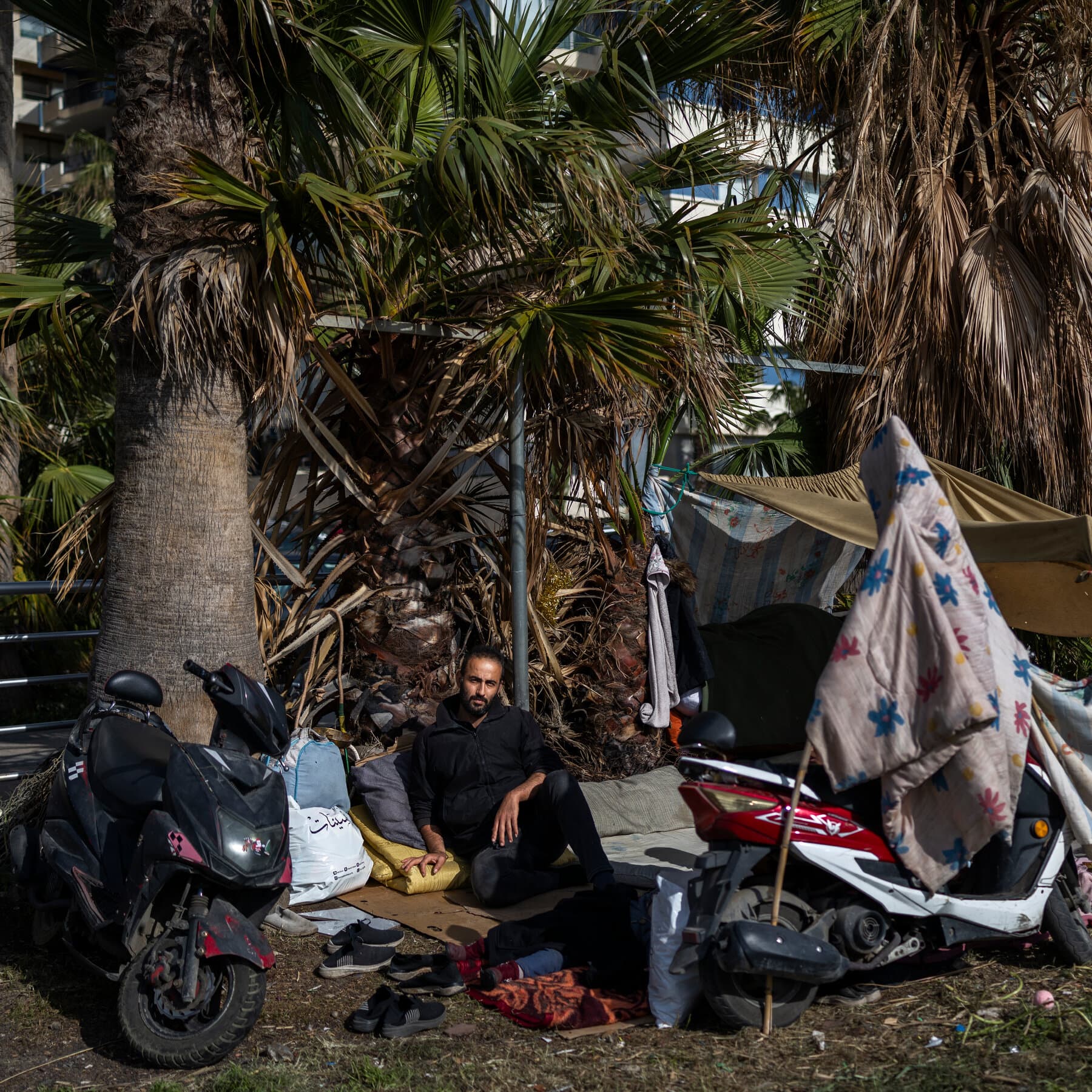 A gritty, hyper-realistic scene at night on a street in Beirut. Families sleeping on thin mattresses on the dirty concrete sidewalk. In the background, smoke rising from a destroyed building skyline, illuminated by orange firelight. The people look exhausted and hopeless. High contrast, cynical atmosphere.