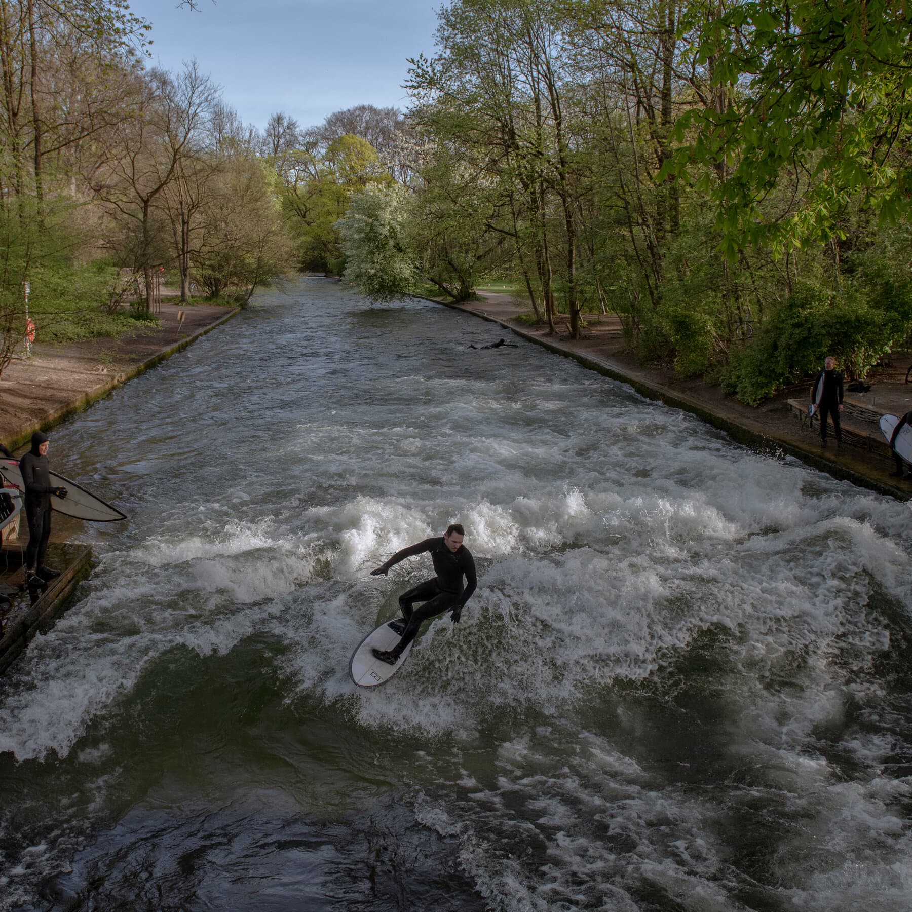 A gloomy, overcast day in an urban park in Munich. A concrete river channel filled with flat, boring grey water. On the concrete bank, a group of three sad men in black neoprene wetsuits are standing and staring at the water with disappointment. They are holding surfboards under their arms. In the background, grey city buildings and bare trees. The mood is depressing and gritty. High contrast.