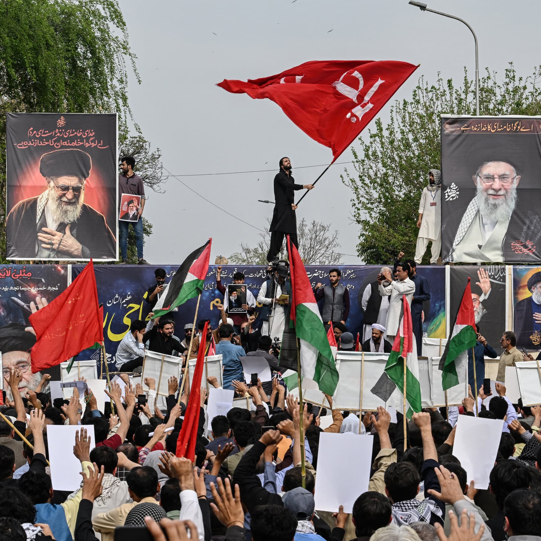 A gritty, high-contrast political illustration showing a darkened map of Pakistan with chaotic riots in the streets, overshadowed by a large, indifferent silhouette of a man in a suit playing with toy missiles on a table, cynical style, dark colors.