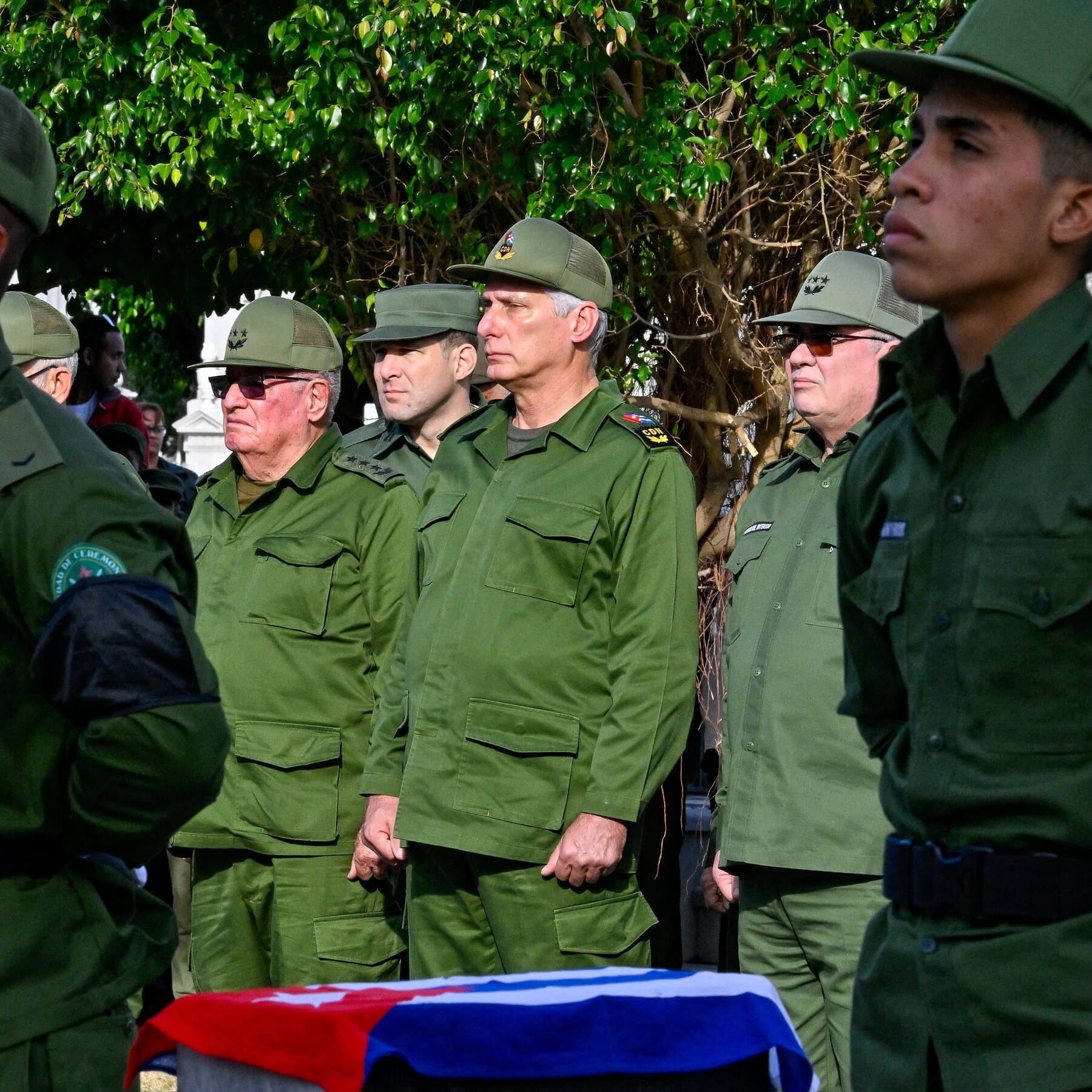 A gritty, cinematic shot of a dark street in Havana at night. A single, flickering yellow streetlamp illuminates a line of old, rusted cars and people standing in a long shadow. In the background, a large, faded propaganda poster of a faceless leader peeling off a wall, while a cold, blue light from the north shines over the horizon like a searchlight.