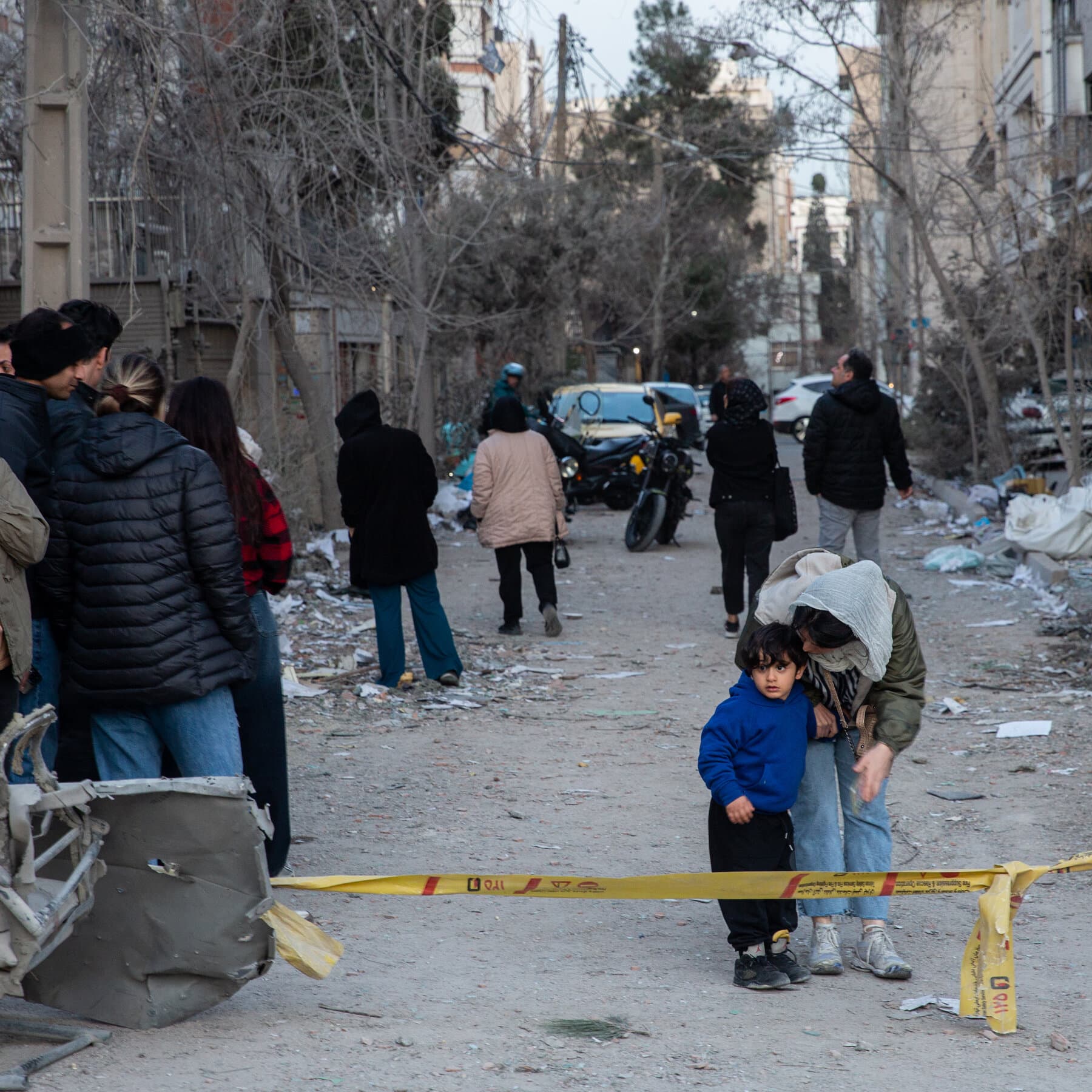 A gritty, high-contrast illustration of a modern city skyline at night under siege. Silhouettes of apartment blocks are visible against a dark sky lit up by distant explosions and streaks of light. In the foreground, the view is from a broken window looking out, emphasizing isolation and fear. The style should be dark, moody, and cynical, focusing on the atmosphere of dread rather than graphic violence.